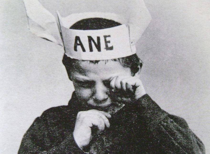 une photo en noir et blanc d'un enfant d'environ 5 ans, qui a sur la tête une couronne de papier avec 2 oreilles et la mention "ANE" . Il pleure et s'essuie les yeux avec sa main repliée.