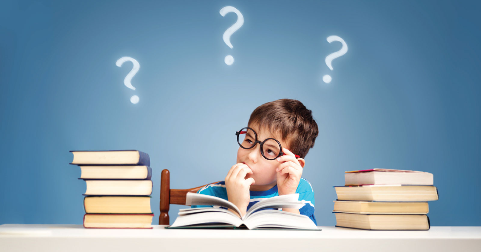 un enfant attablé à un bureau se pose des questions au sujet d'un livre ouvert devant lui. Sur le bureau 2 piles de livre, une à gauche l'autre à droite. En fond, un mur bleu sur lequel sont 3 points d'interrogation