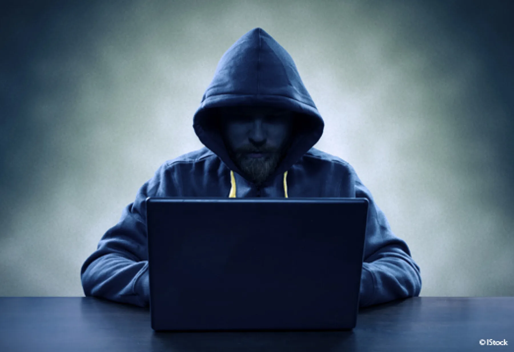 un homme assis à un bureau devant un ordinateur. Son visage est à peine visible sous la capuche d'une veste de couleur gris foncé, et il est dans la pénombre. La photo est grise et sombre.