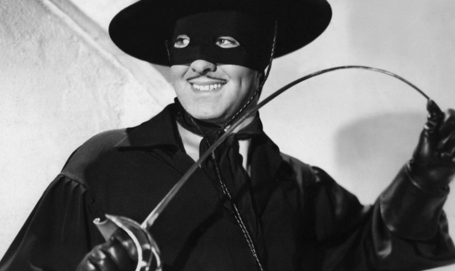 Tyrone Power dans le rôle de Zorro : habillé en noir, un masque noir lui couvrant le haut du visage à l'exception des yeux, un chapeau noir, une chemise noire et des gants noirs, souriant et tenant entre les mains une épée qu'il recourbe.